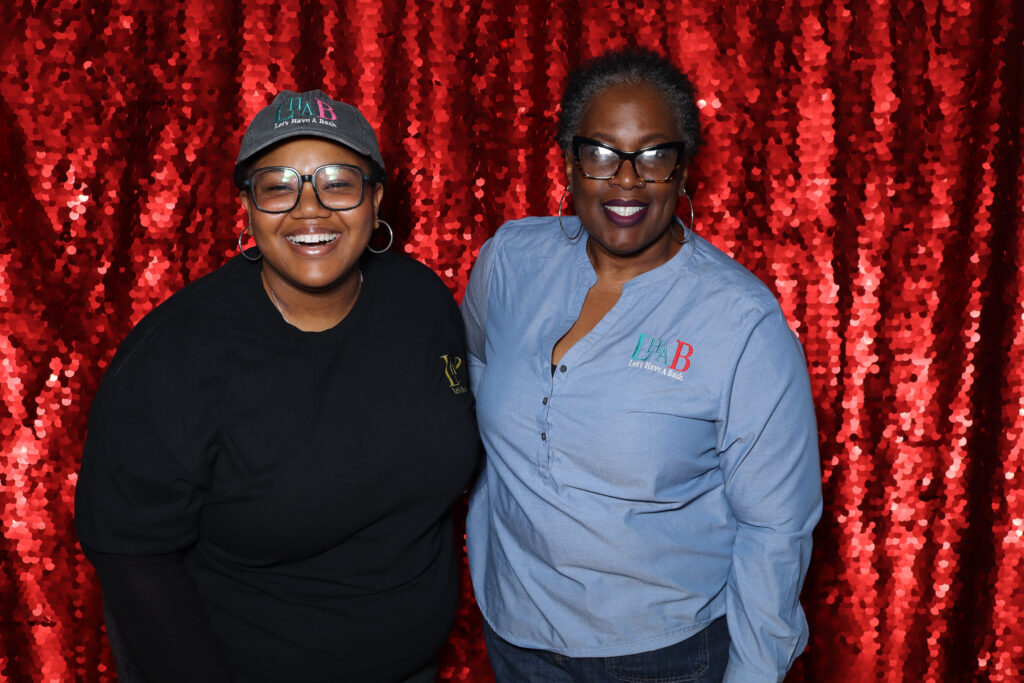 IMG_8268 two black women standing in front of a red backdrop. One woman is wearing a black shirt, the other woman is wearing a blue buttondown shirt